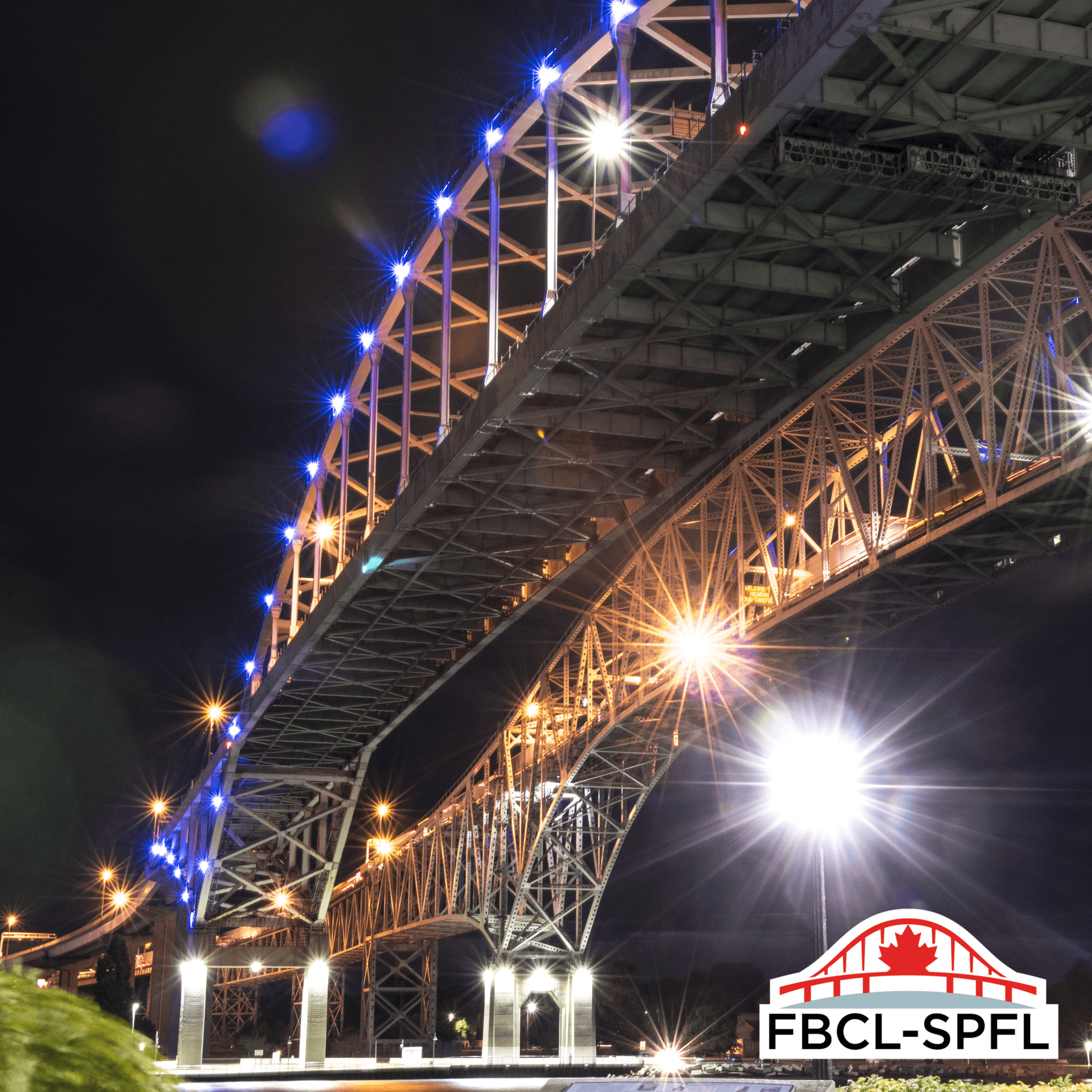 Vue nocturne en contre-plongée sous les deux travées du pont Blue Water. Des lumières DEL bleues illuminent la travée supérieure, tandis que des lumières blanc chaud éclairent la travée en dessous. Le logo de la SPFL apparaît dans le coin inférieur droit.